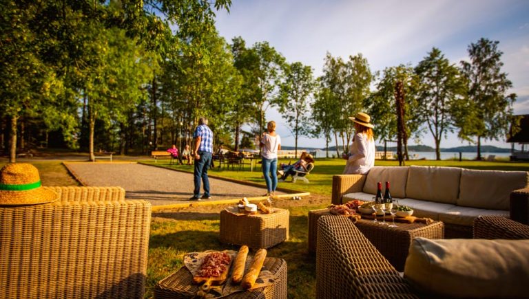 Midsommarfirande på gräset med boule och lekar. I förgrunden finns loungemöbler och i bakgrunden syns människor, några spelar boule.