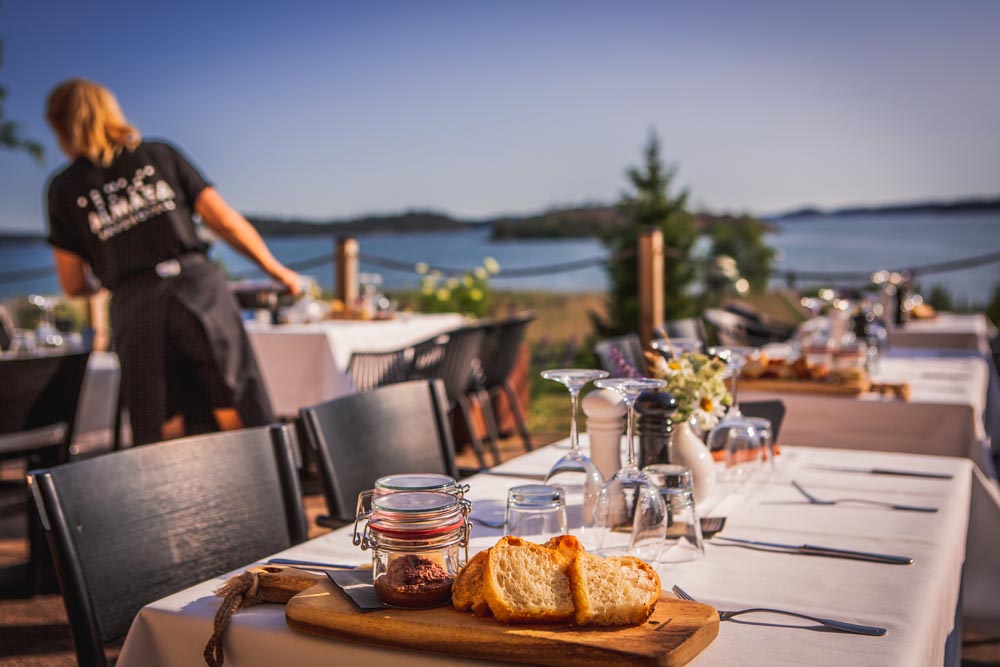 Sommar på terrassen, i förgrunden dukade bord med bröd och tapenad. Till vänster dukar Tina fler bord och i bakgrunden ligger Hårsfjärden alldeles blå. 