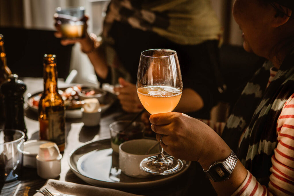 Bilden föreställer en hand som håller i en måltidsdryck, bakgrunden är bord med personer i ofokus som njuter av Almåsas brunch