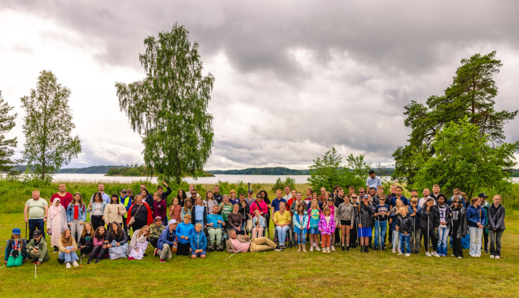 Gruppbild på Unga med synnedsättning samt barn från SRF's barnläger på plats på gräsmattan med Hårsfjärden som bakgrund.